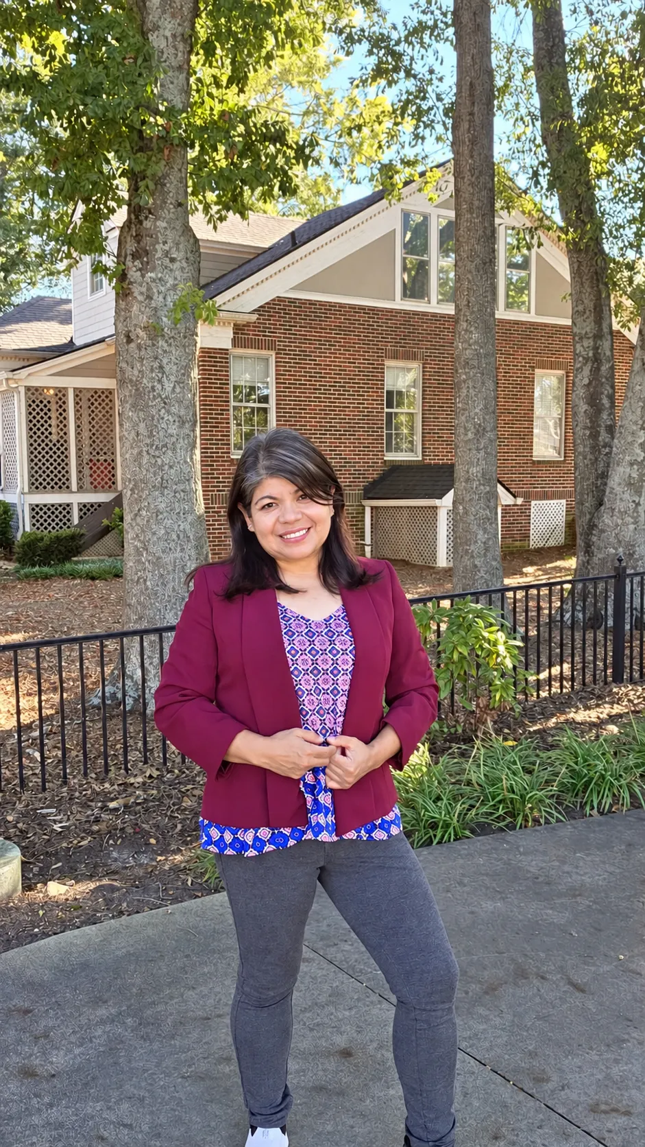 Erika Rubio in a burgundy blazer, standing on a tree-lined sidewalk in front of a brick home